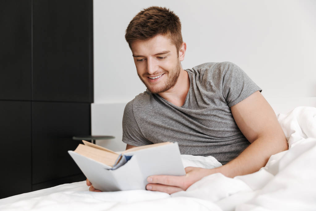 Usměvavý mladý muž s knihou a ležím v posteli doma - Fotografie, Obrázek