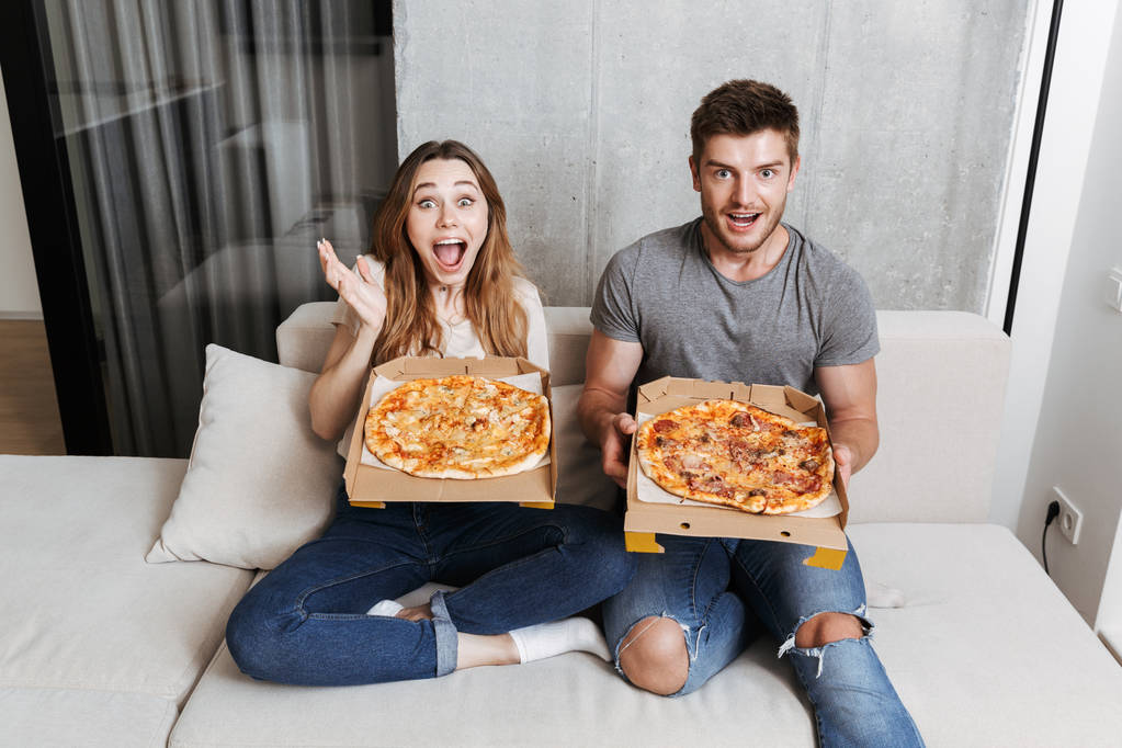 Веселая молодая пара держит коробки с пиццей, сидя вместе на диване дома
 - Фото, изображение