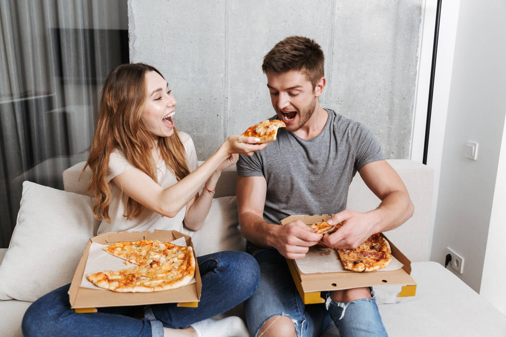 Uśmiechnięta Młoda para jedzenie pizzy razem siedząc na kanapie w domu - Zdjęcie, obraz