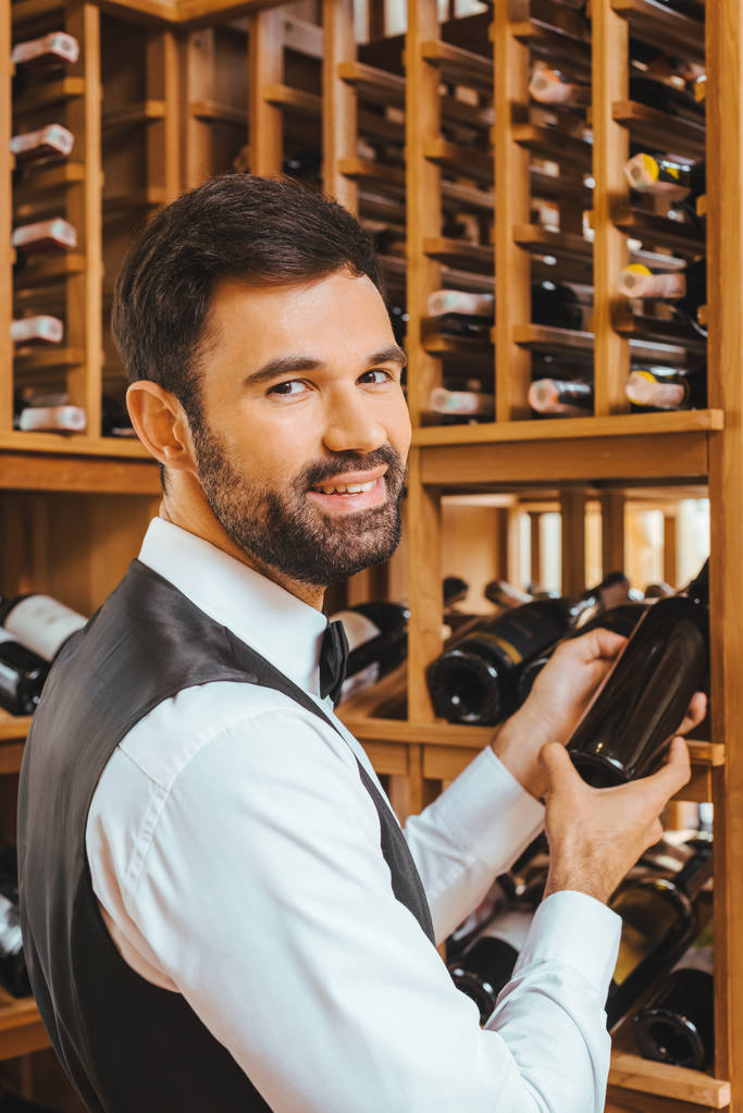 guapo joven sommelier tomando botella de estante en la tienda de vinos y mirando a la cámara
 - Foto, Imagen