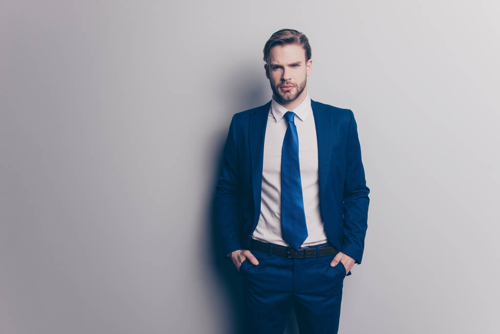 Portret van prachtige, aantrekkelijke, sexy, brutale man in blauwe pak met stropdas met stoppels, houden twee handen in de zakken van de broek, op zoek naar ta camera, geïsoleerd op een grijze achtergrond - Foto, afbeelding