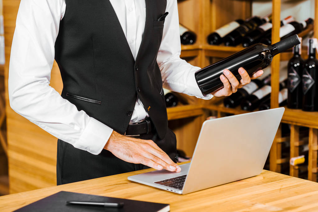 cropped shot of wine steward holding bottle and working with laptop at wine store - Photo, Image