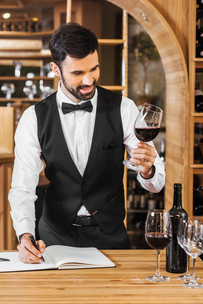 smiling young sommelier examining color of wine and making notes at wine store - Photo, Image