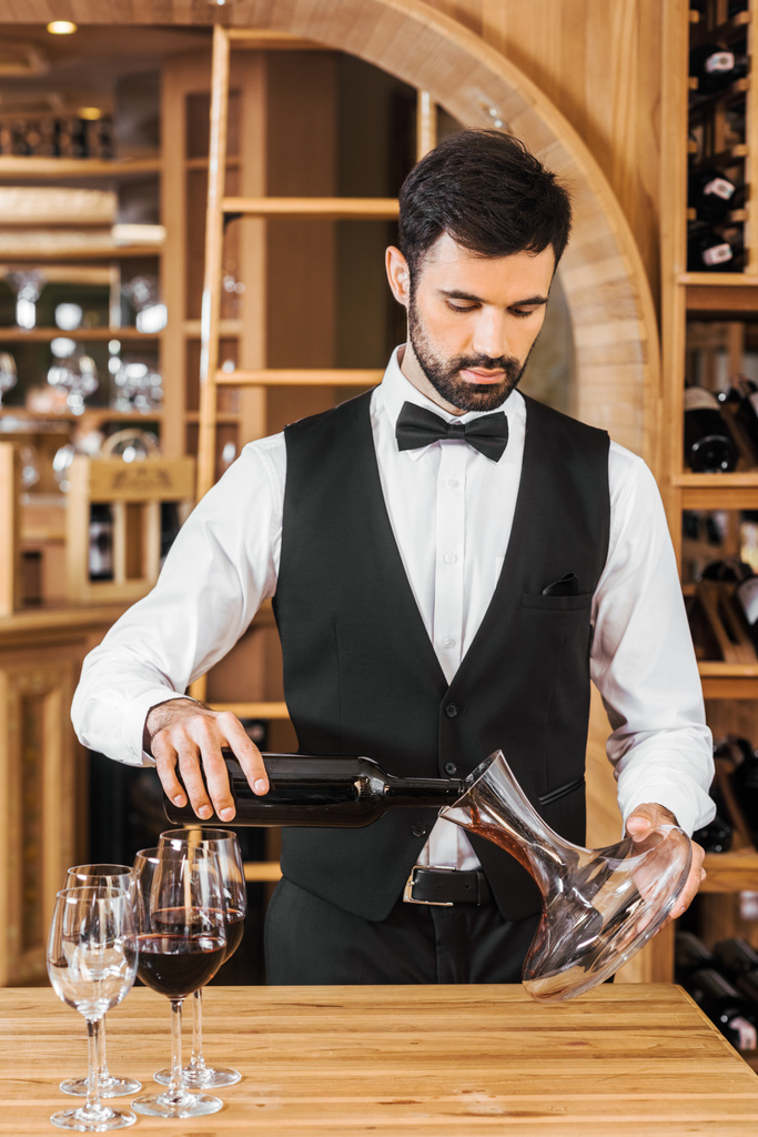 handsome young wine steward pouring wine into decanter at wine store - Photo, Image