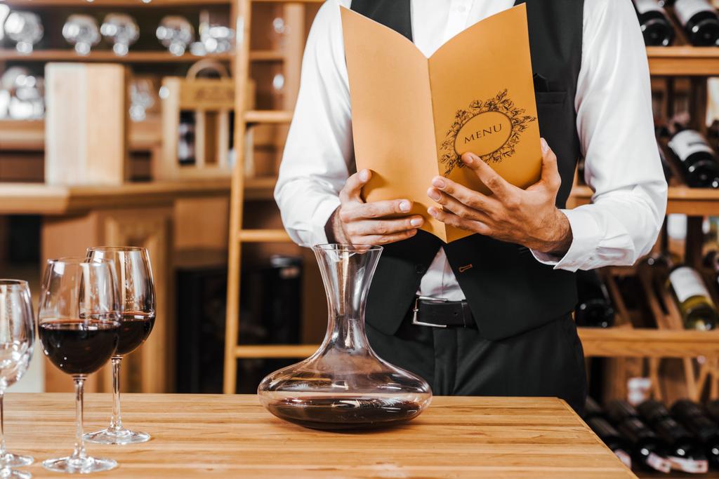 tiro recortado de sommelier con tarjeta de vino de pie delante de la mesa con decantador y copas en la tienda de vinos
 - Foto, Imagen
