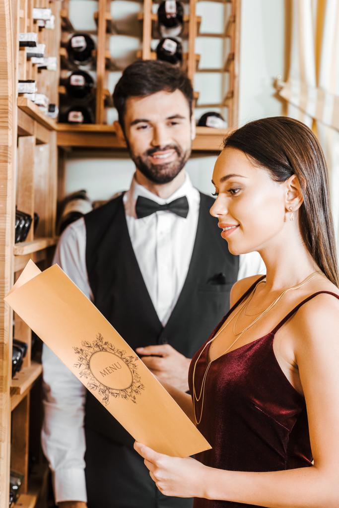 mulher sorrindo segurando cartão de menu na loja de vinho enquanto mordomo de pé embaçado no fundo
 - Foto, Imagem