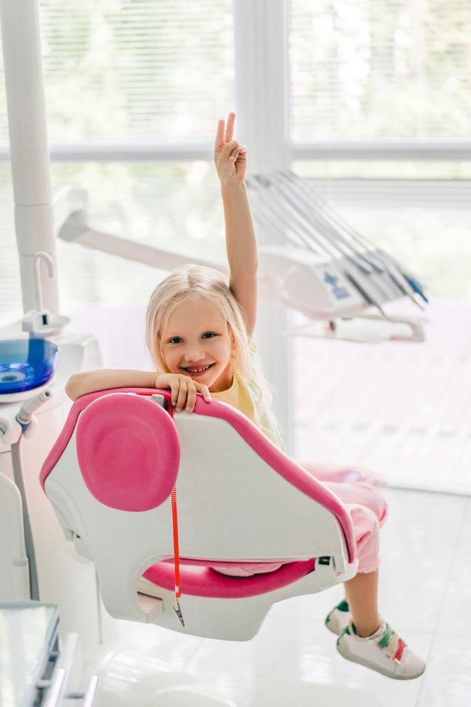 niño sonriente mostrando señal de paz mientras está sentado en la silla en la oficina del dentista
 - Foto, Imagen