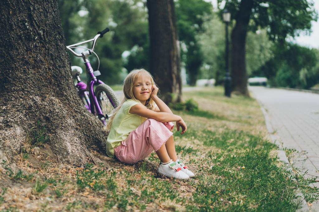Zijaanzicht van de schattige lachende kind rusten in de buurt van fiets onder boom in het park - Foto, afbeelding