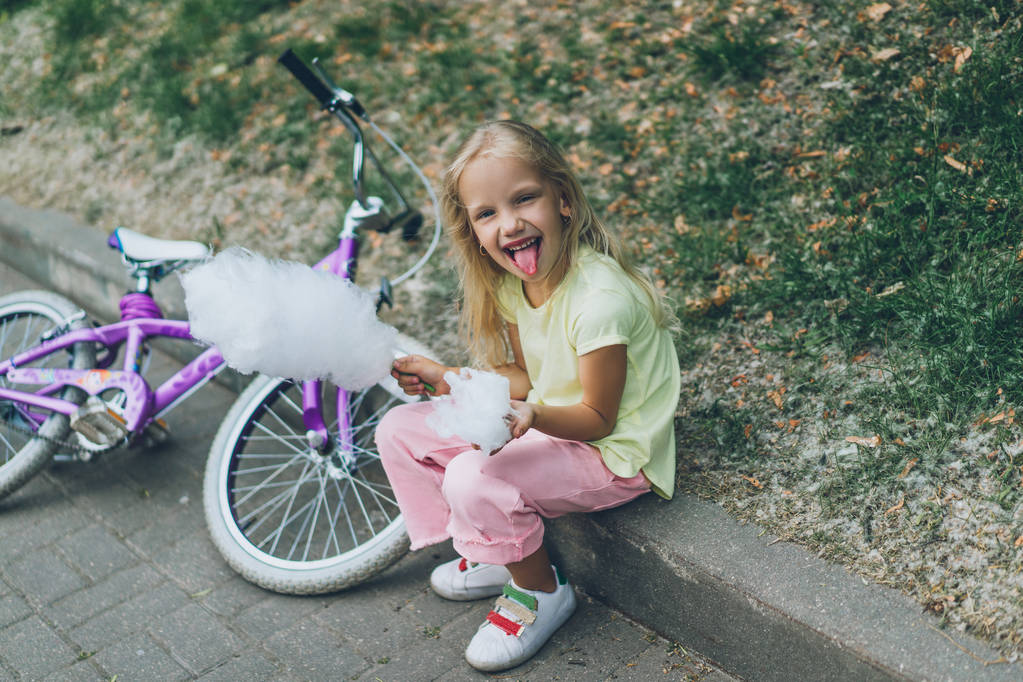 cheerful kid with cotton candy sticking tongue out while sitting on border in park - Photo, Image