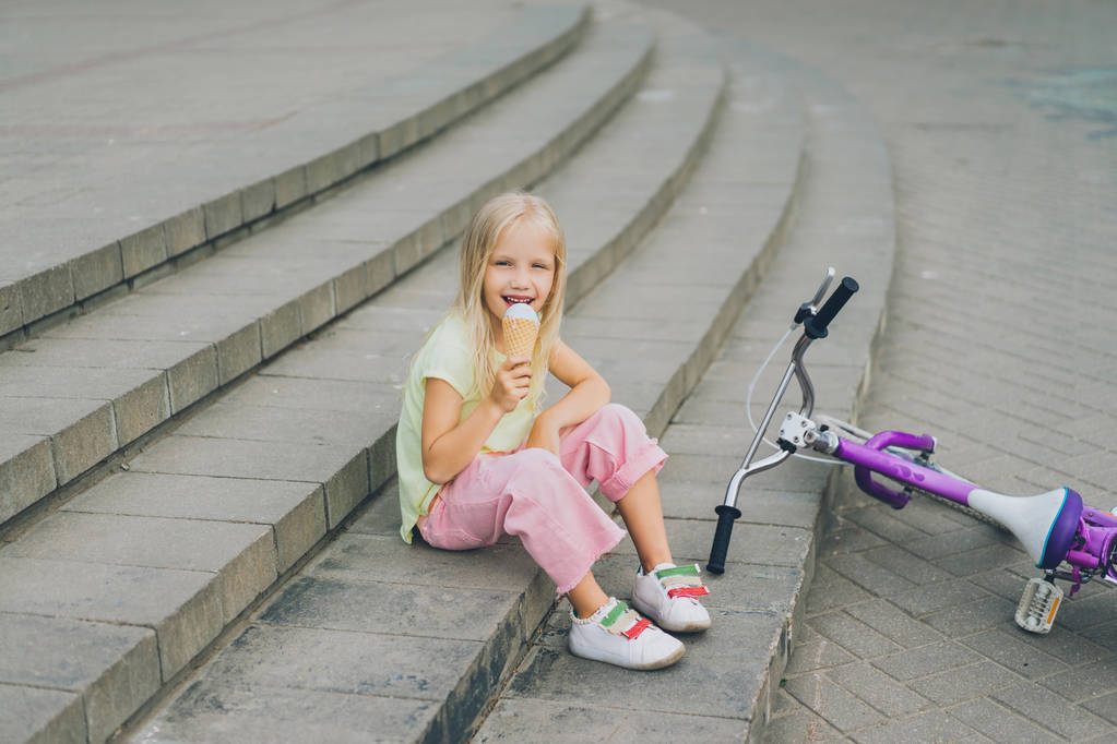 Niedliches Kind isst Eis, während es allein auf der Stadttreppe neben dem Fahrrad sitzt - Foto, Bild