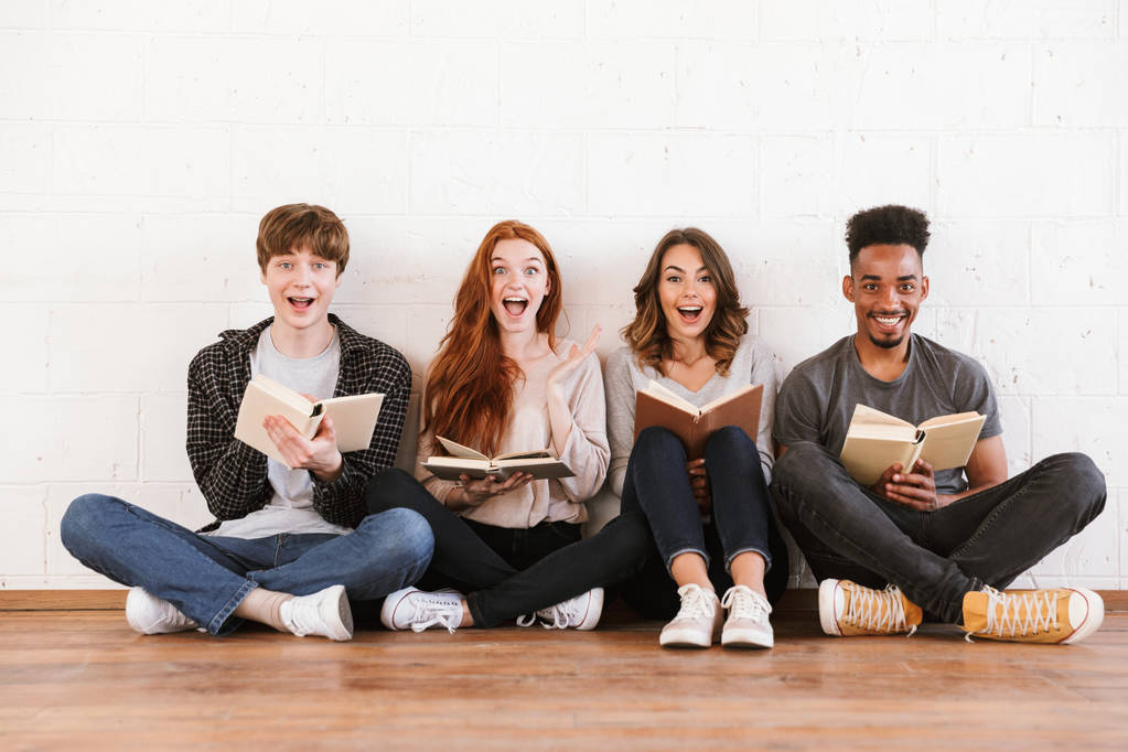 Bild von überraschten emotionalen jungen Freunden Studenten, die über weißen Wandhintergrund sitzen und drinnen vor der Kamera Bücher lesen. - Foto, Bild
