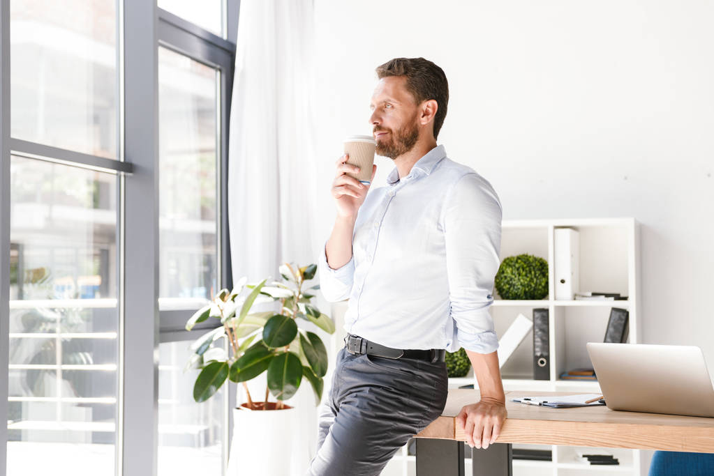 Hombre de negocios sonriente bebiendo café mientras se apoya en un escritorio en la oficina
 - Foto, Imagen