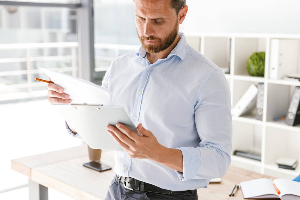 Imagen recortada del hombre de negocios analizando documentos mientras está de pie y apoyado en un escritorio en la oficina
 - Foto, imagen