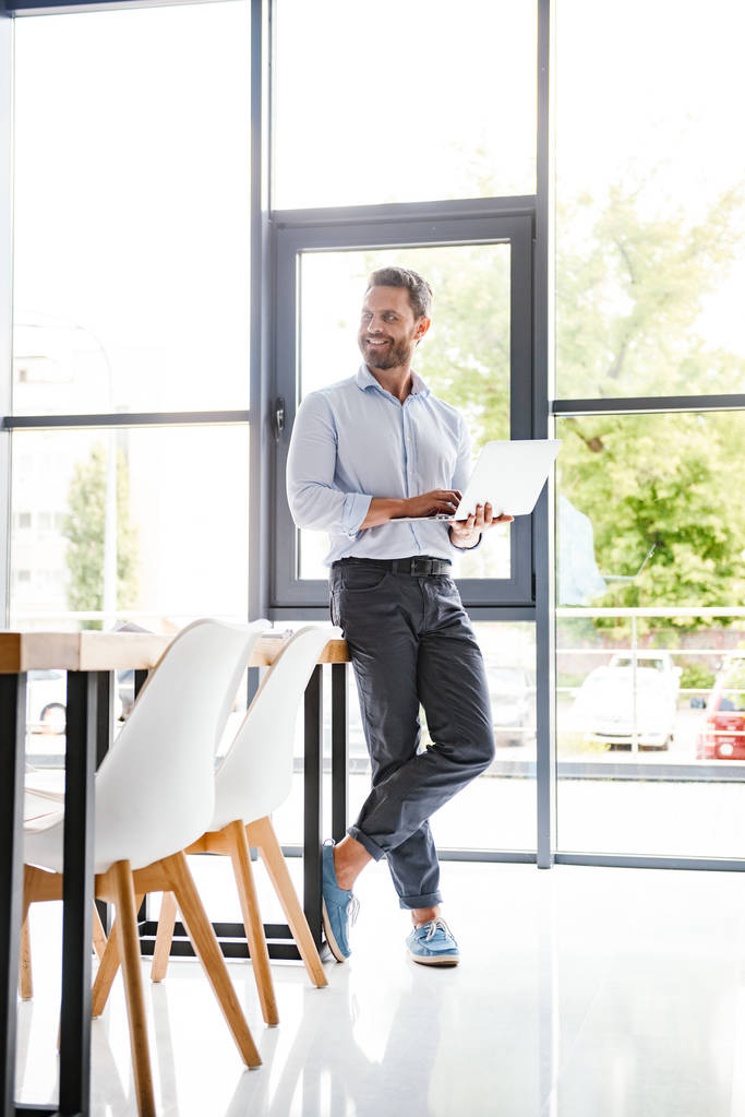 Image de l'homme barbu heureux dans le bureau debout à l'intérieur en utilisant un ordinateur portable travaillant en regardant de côté
. - Photo, image