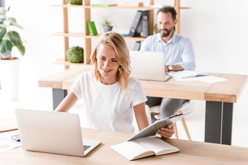 Mujer sonriente gerente que trabaja en el ordenador portátil mientras está sentado en la oficina con el hombre compañero de trabajo en un segundo plano
 - Foto, Imagen