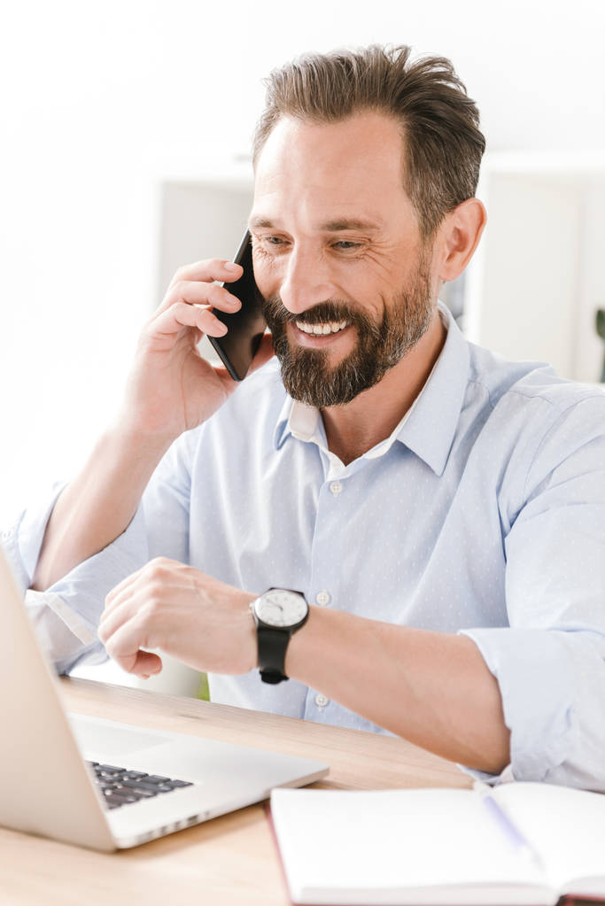 Счастливый бизнесмен разговаривает по мобильному телефону, сидя за рабочим столом с бумагами и ноутбуком
 - Фото, изображение