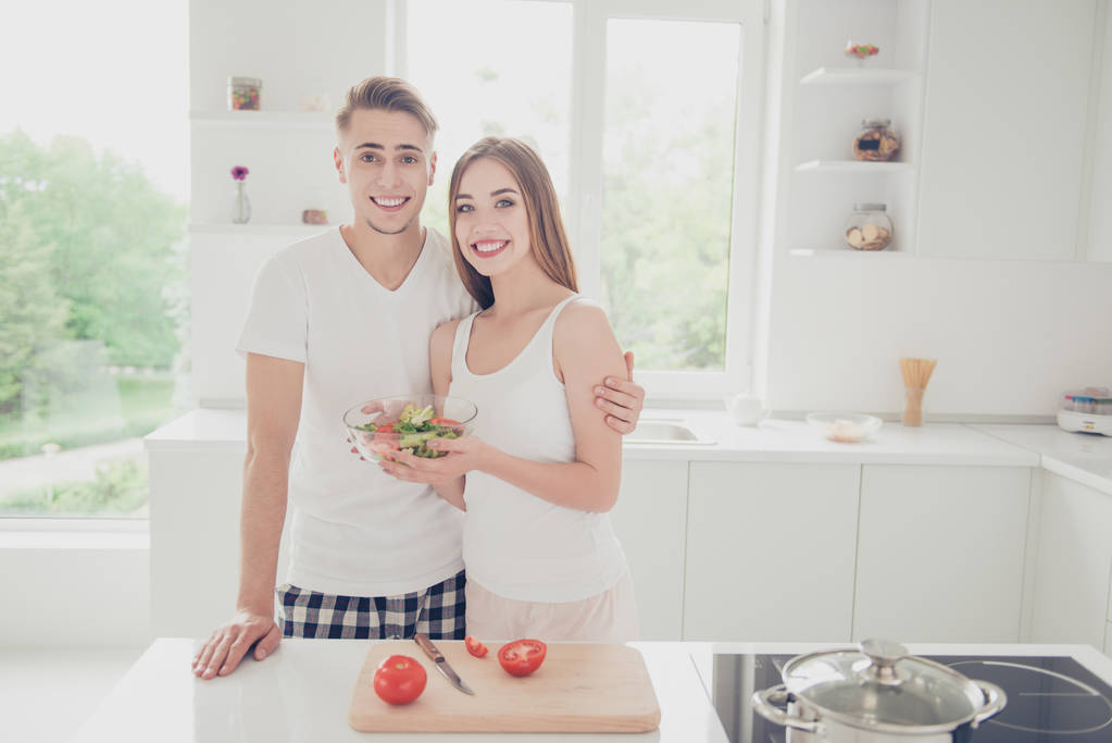 Портрет молодой привлекательной милой милой пары вместе на светлой кухне, носить пижаму, приготовление пищи, салат, показывая готовое блюдо в миске, улыбаясь
 - Фото, изображение