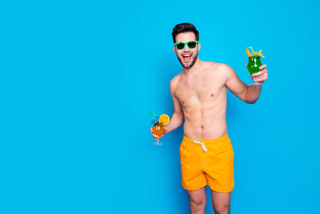 Пляжний стиль! Привабливий і веселий чоловік в жовтих шортах і окулярах, тримає коктейлі з невеликою парасолькою в руках, ізольовані на синьому фоні. Концепція радісного гарячого літа
 - Фото, зображення