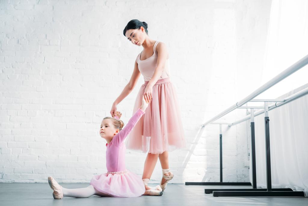 young ballet teacher looking at child in pink tutu stretching in ballet school - Photo, Image