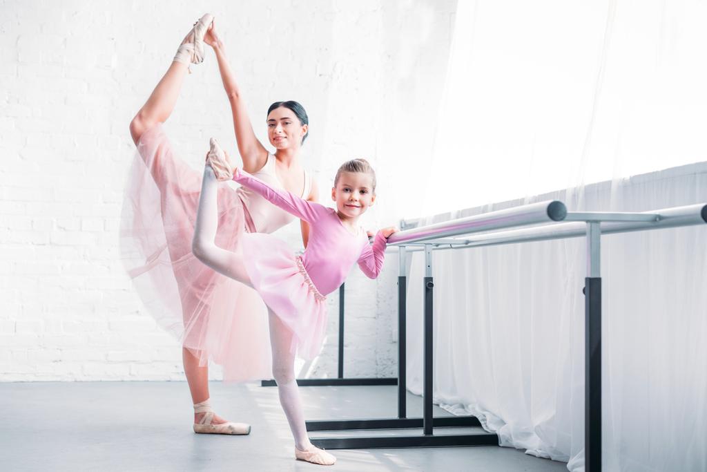 beautiful adult and little ballerinas stretching and smiling at camera in ballet school - Photo, Image