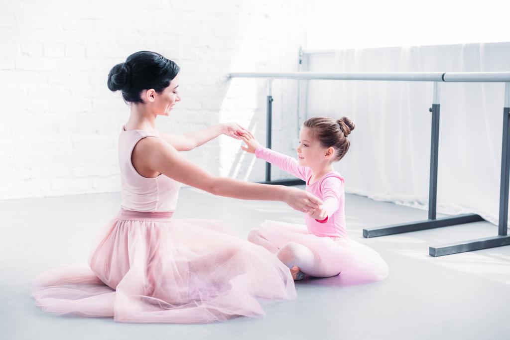 Zijaanzicht van de ballet leraar en beetje student in roze tutu rokken zittend en bedrijf handen tijdens een training in de balletschool - Foto, afbeelding