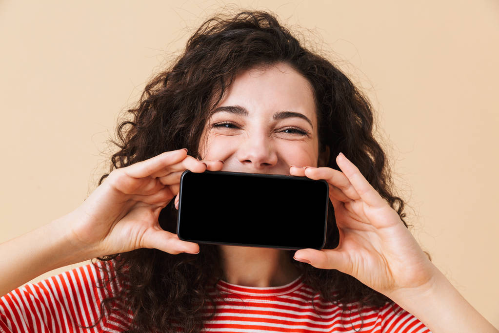 Εικόνα του χαριτωμένο σγουρά νεαρή γυναίκα που στέκεται απομονωμένο πάνω μπεζ φόντο οθόνης από κινητό τηλέφωνο που καλύπτουν το στόμα. - Φωτογραφία, εικόνα