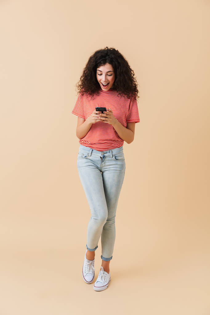 Ganzkörperporträt eines glücklichen, lässigen Mädchens mit Mobiltelefon auf beigem Hintergrund - Foto, Bild