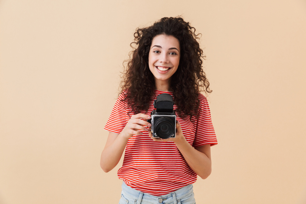 Fotoğraf kamera tutarak bej renkli arka plan üzerinde izole güzel genç kıvırcık kadın fotoğrafçı. - Fotoğraf, Görsel