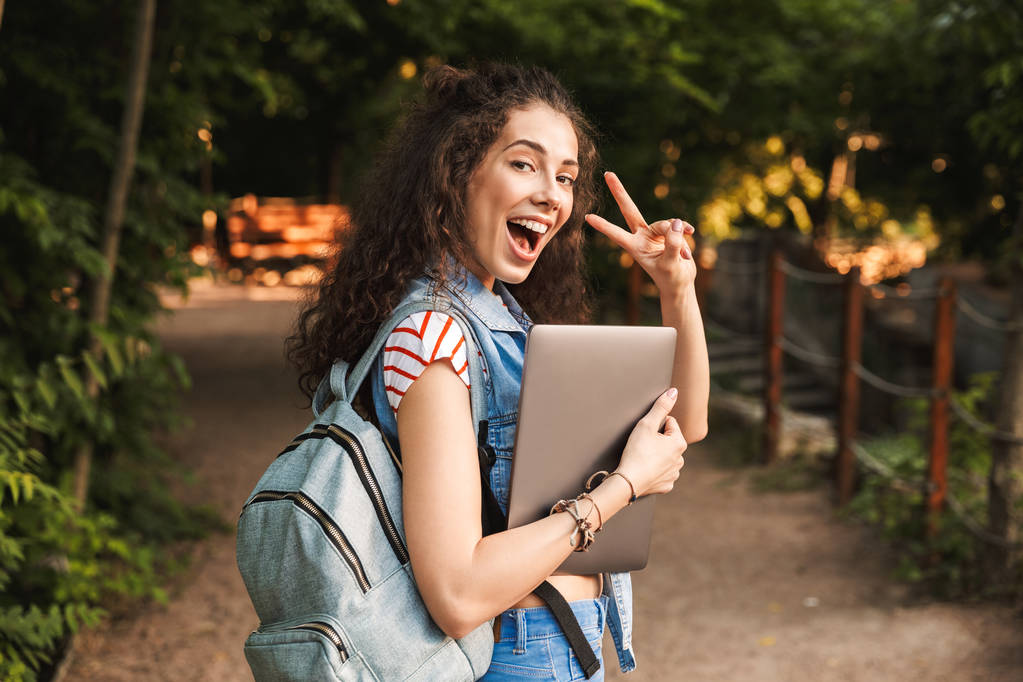 Snímek šťastné mladé krásné ženy držící přenosný počítač ukazuje míru gesto chůzi venku v parku. Při pohledu kamery. - Fotografie, Obrázek