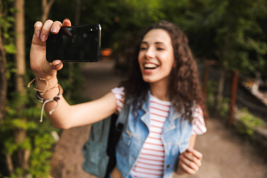 Wazige foto van mooie brunette vrouw 18-20 met rugzak breed glimlachend en nemen selfie foto op mobiele telefoon tijdens het wandelen langs de weg in groen park - Foto, afbeelding