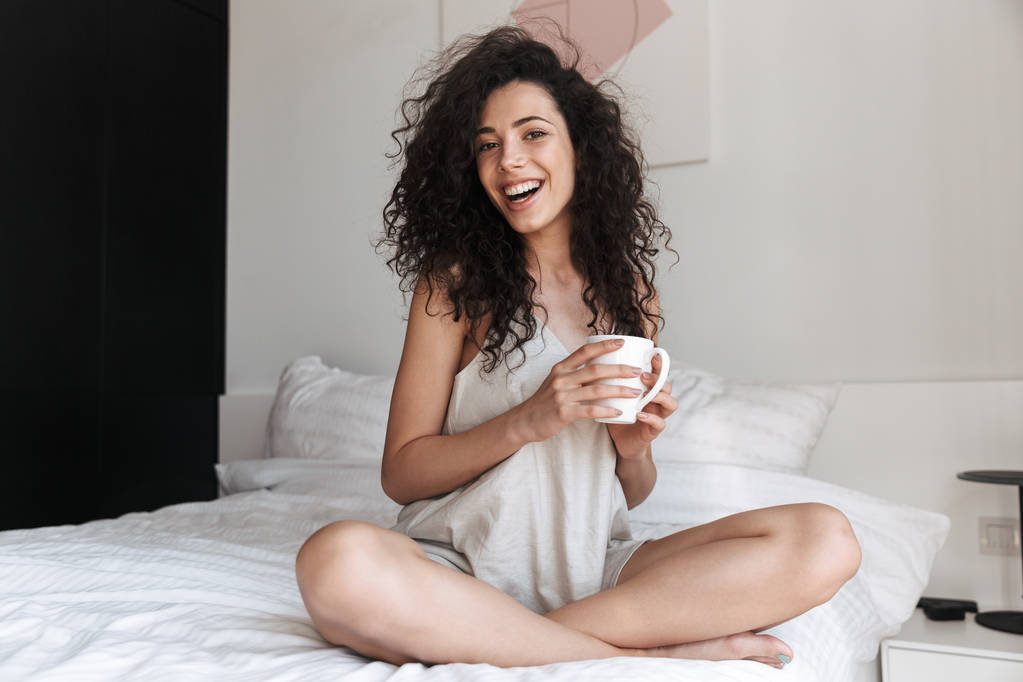 Ritratto di splendida donna felice con lunghi capelli ricci seduta sul letto con biancheria bianca pulita a casa al mattino e con in mano una tazza di tè
 - Foto, immagini