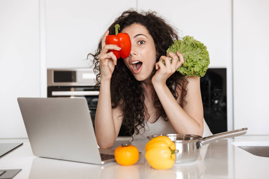 Foto van krullend vrouw 20s groenten minnaar groene salat en zoete papier te houden tijdens het koken diner in keuken en lezing recept van laptop - Foto, afbeelding