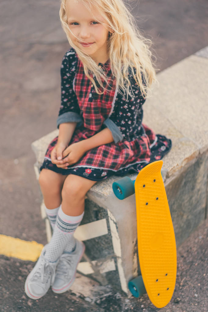 adorável sorrindo criança sentado perto de skate e olhando para a câmera na rua urbana
 - Foto, Imagem