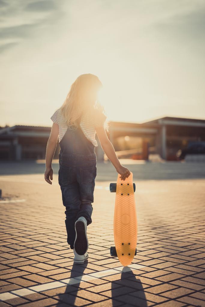 rear view of little kid walking with skateboard at parking lot with setting sun behind  - Photo, Image