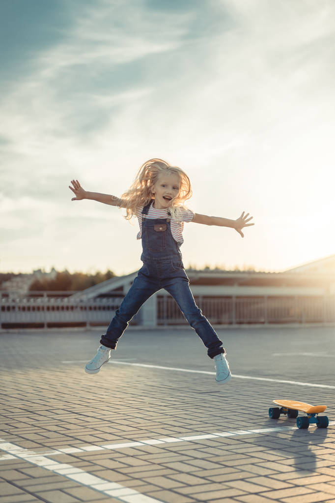 adorable little child jumping with wide arms near skateboard at parking lot  - Photo, Image