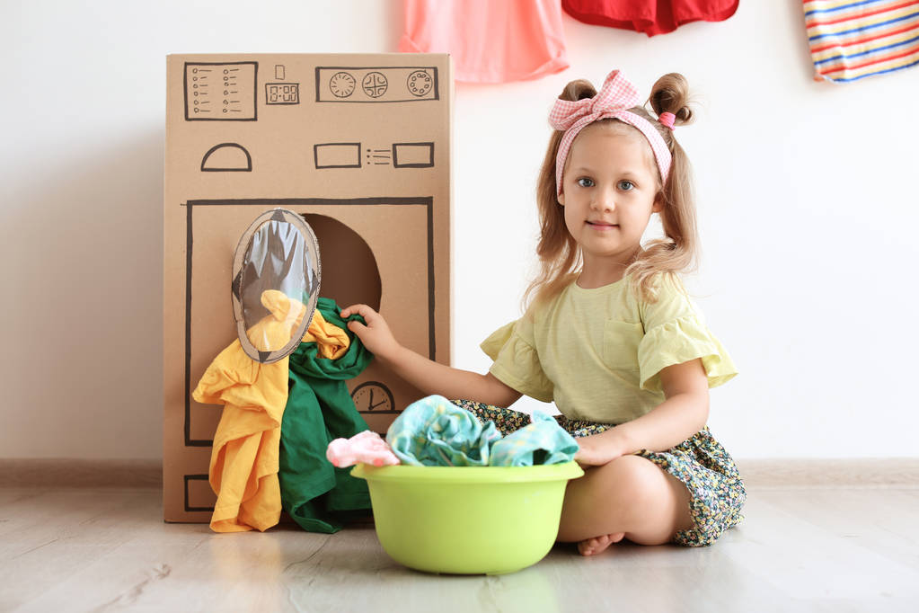 Suloinen pieni lapsi leikkii pahvin pesukoneella ja vaatteilla sisätiloissa
 - Valokuva, kuva