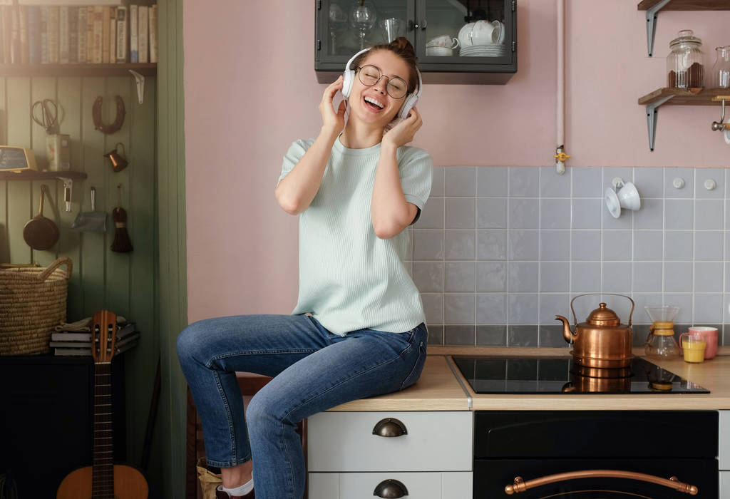 Foto orizzontale della donna caucasica europea che si gode il tempo libero nel suo appartamento usando parte del tavolo della cucina come sedile, muovendosi e sorridendo mentre sta ascoltando tracce audio positive nel suo lettore
 - Foto, immagini