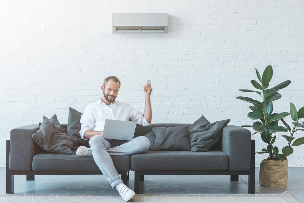 Mann schaltet Klimaanlage mit Fernbedienung ein, während er Laptop auf Sofa benutzt - Foto, Bild