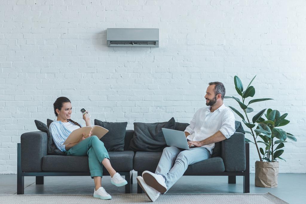 couple allumant le climatiseur pendant la chaleur estivale tout en étant assis sur le canapé avec livre et ordinateur portable
  - Photo, image