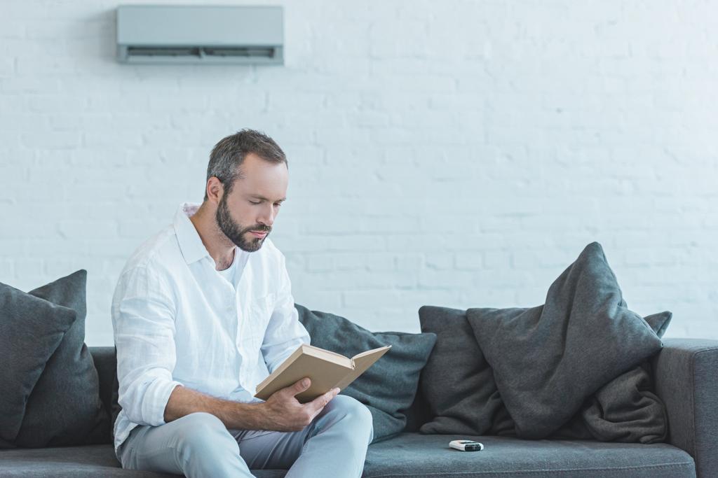 homem barbudo livro de leitura no sofá, com ar condicionado na parede
 - Foto, Imagem