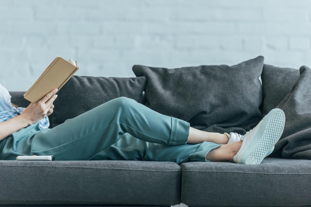 περικοπεί δείτε γυναίκα χαλαρώνοντας στον καναπέ και την ανάγνωση του βιβλίου  - Φωτογραφία, εικόνα