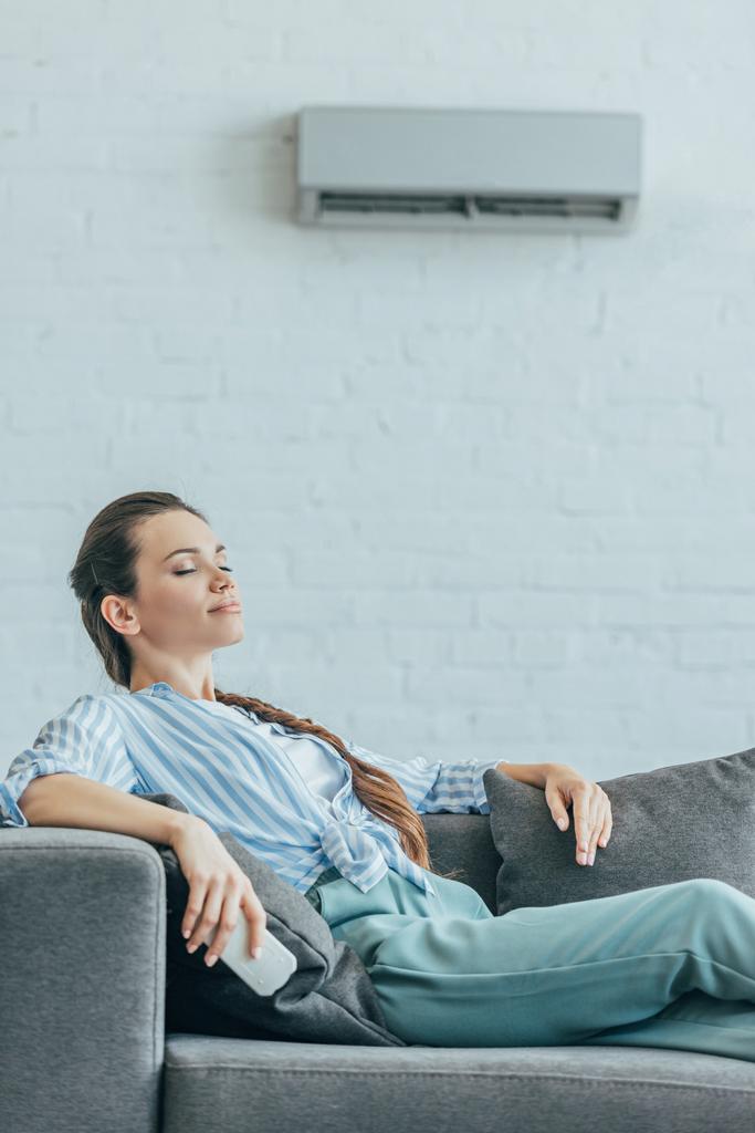 woman resting on sofa with air conditioner on wall, summer heat concept - Photo, Image
