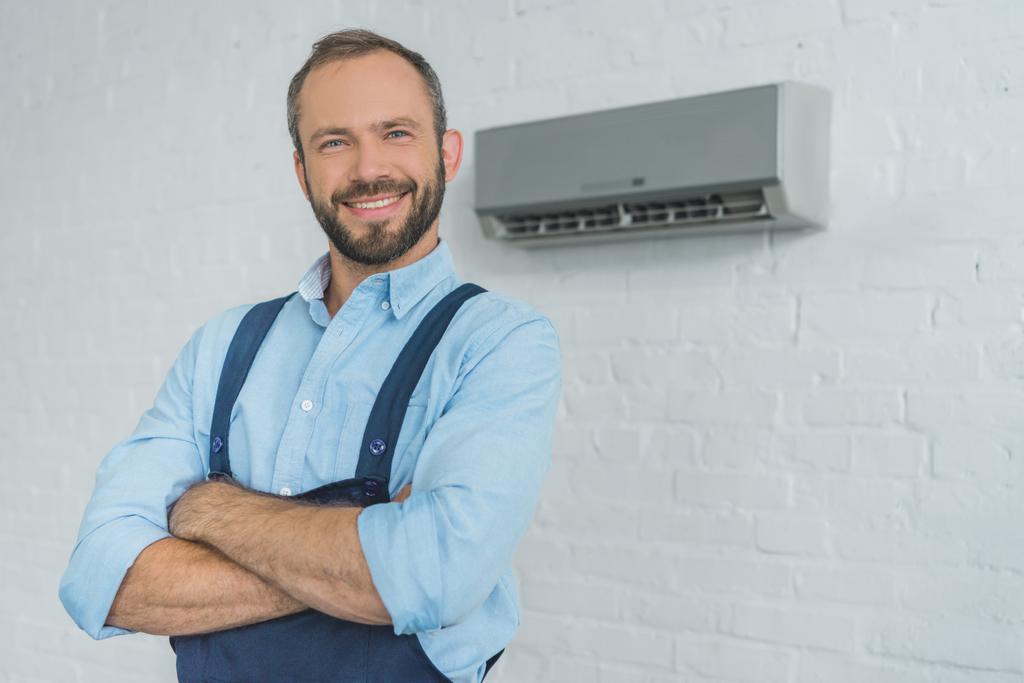 glimlachend bebaarde reparateur poseren met gekruiste armen, met air conditioner op muur - Foto, afbeelding