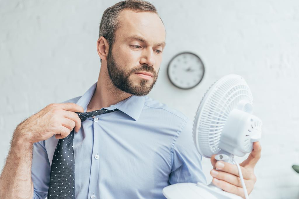 zakenman verwijderen stropdas en blazen op zichzelf met witte elektrische ventilator   - Foto, afbeelding