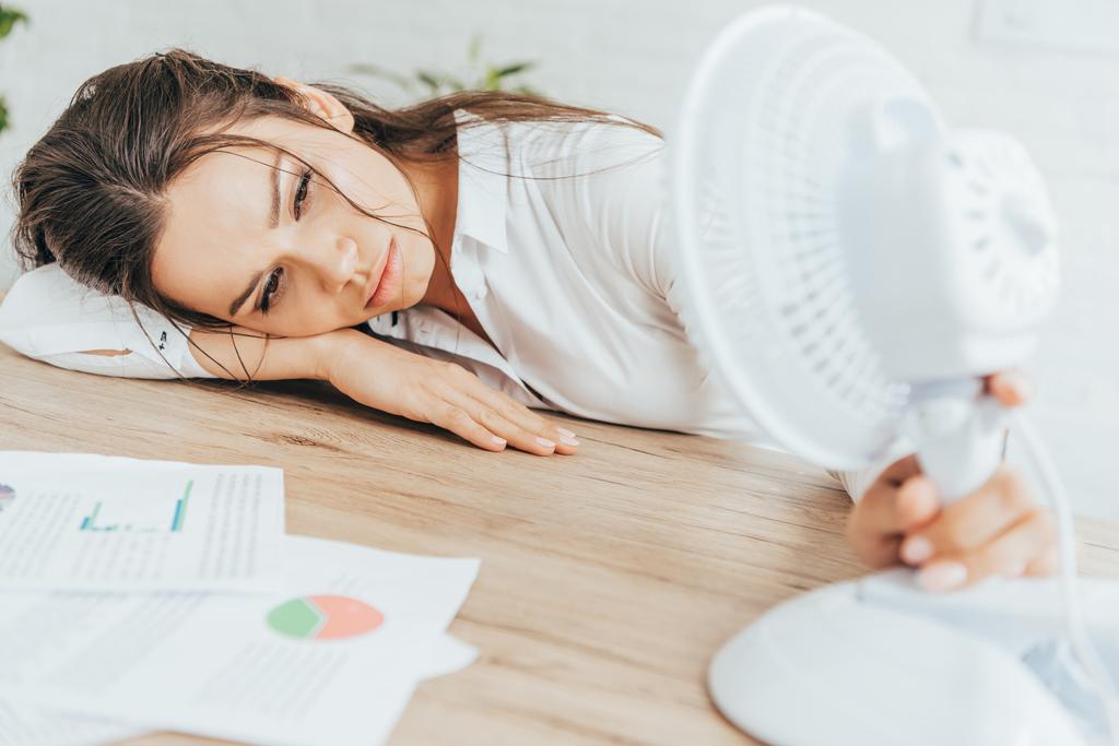 mulher de negócios exausta soprando em si mesma com ventilador elétrico enquanto deitada na mesa com documentos
 - Foto, Imagem