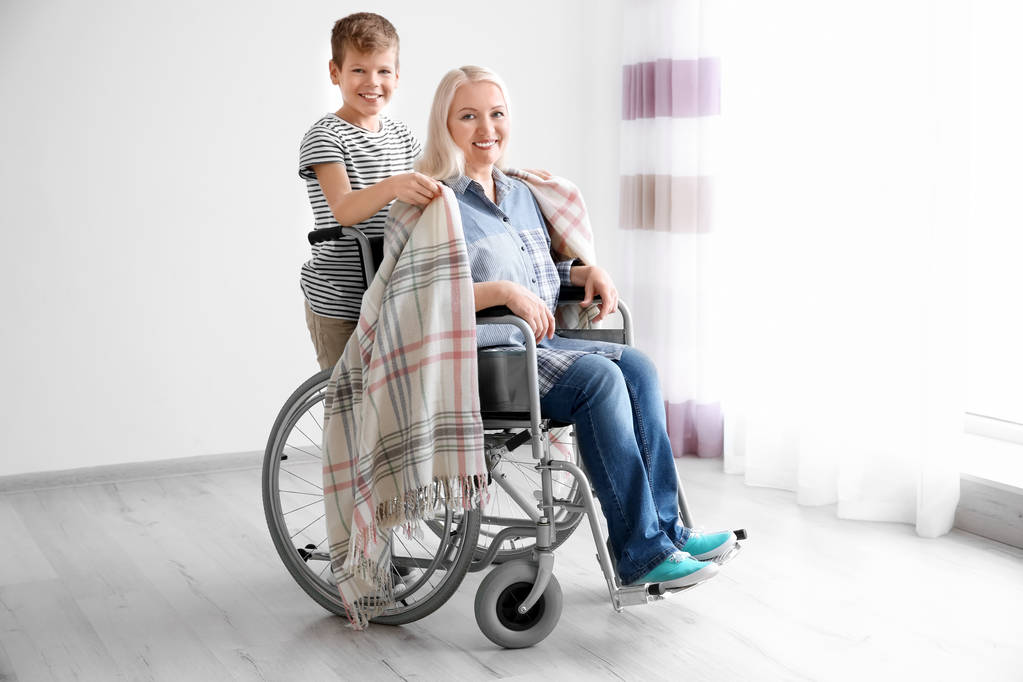 Kleine jongen verzorgen van rijpe vrouw in rolstoel binnenshuis - Foto, afbeelding