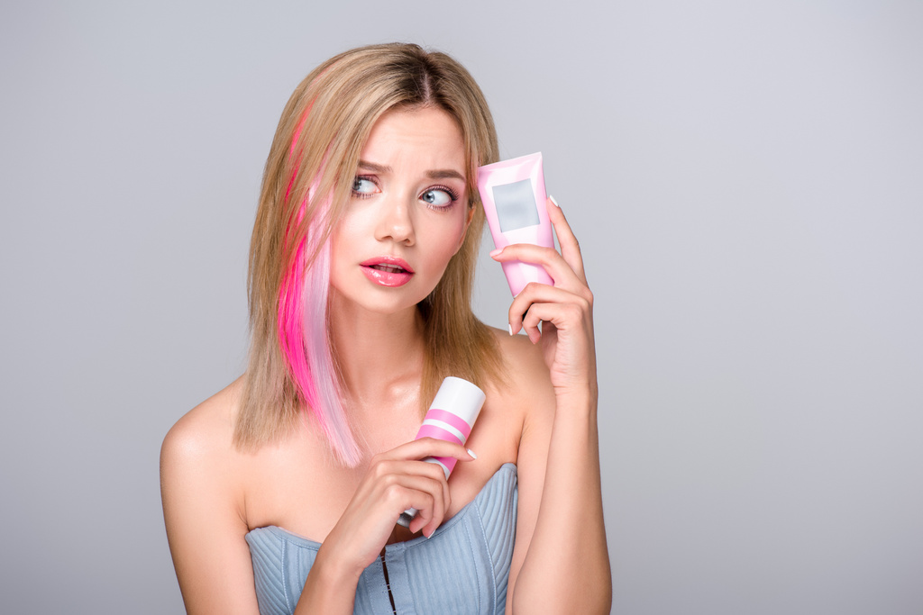 bewildered young woman with colored bob cut holding hair care supplies isolated on grey - Photo, Image