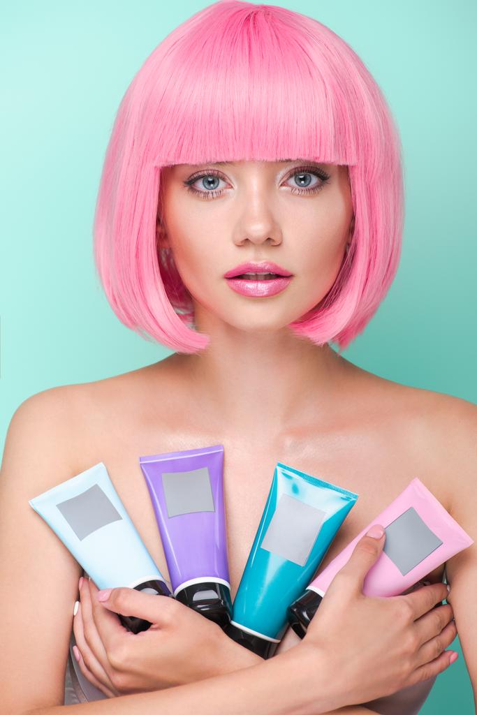attraente giovane donna con taglio di bob rosa in possesso di vari tubi di colorare i tonici dei capelli guardando la fotocamera isolata su turchese
 - Foto, immagini
