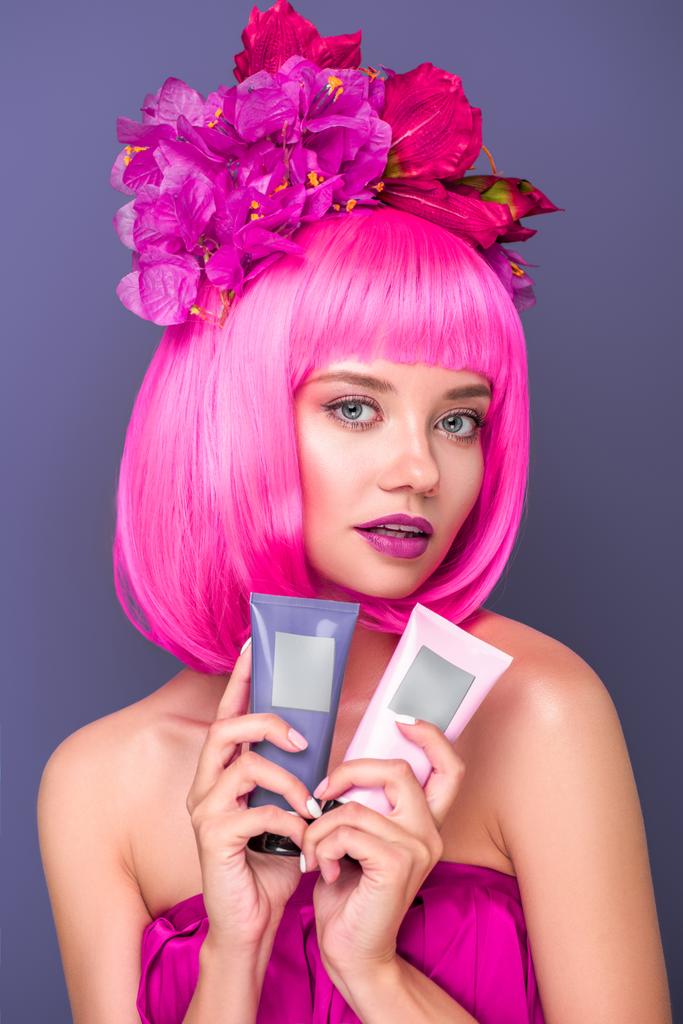 beautiful young woman with pink bob cut and flowers in hair holding tubes of coloring hair tonics isolated on violet - Photo, Image