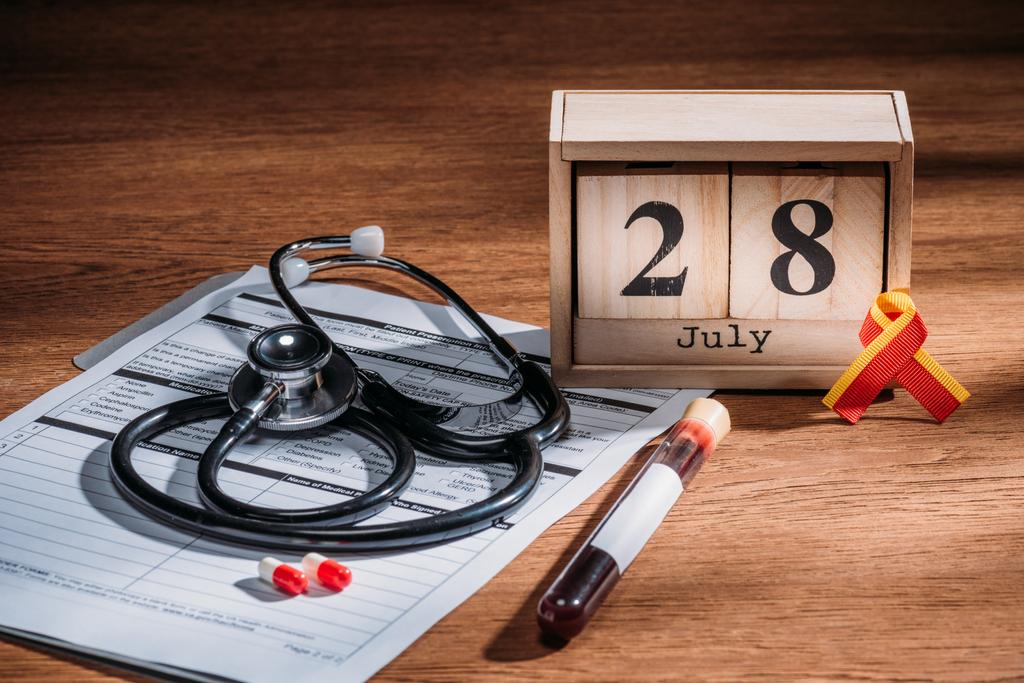 селективный фокус деревянного календаря с 28-й июльской датой, медицинская анкета, таблетки, стетоскоп, колба с образцом крови, концепция дня гепатита в мире
 - Фото, изображение
